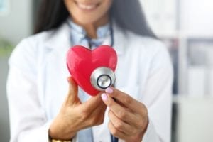 Hands of female GP holding stethoscope head near red toy heart