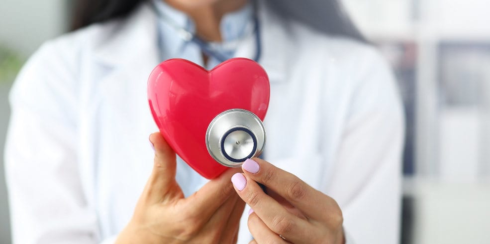 Hands of female GP holding stethoscope head near red toy heart Hands of female GP holding stethoscope head near red toy heart